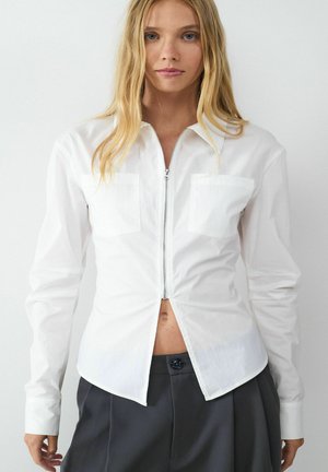 Weißes langärmliges Hemd mit einem Reißverschluss auf der Vorderseite, zwei Fronttaschen, abgerundetem Saum und einer glatten Textur. Kombiniert mit grauen Hosen.