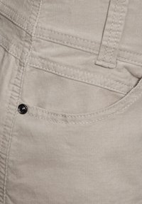 Vaalean beige corduroy-kangas, jossa on sileä pinta, valkoisia ompeleita ja musta nappi taskussa. Lähikuvassa housujen yksityiskohta.