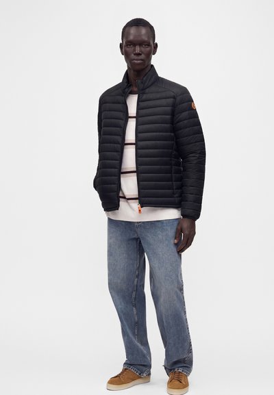 Modèle masculin portant une veste matelassée noire, une chemise blanche rayée, un jean bleu et des baskets marron, debout devant un fond uni.
