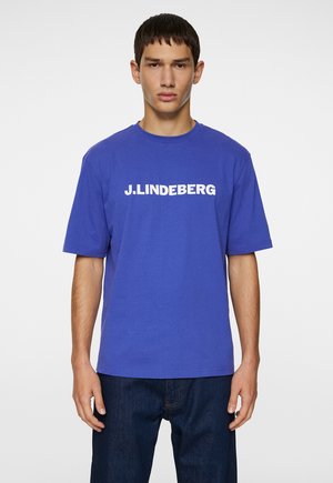 Blå bomulds T-shirt med korte ærmer har et stort hvidt "J. LINDEBERG" logo på brystet. Enkel rund halsudskæring og afslappet pasform.