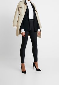 Beige Trenchcoat über einem schwarzen Blazer und figurbetonten schwarzen Hosen. Es sind hohe Absätze und lange Ärmel vorhanden, die einen geschichteten Kleidungseffekt erzeugen.