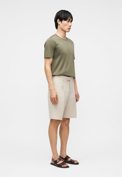 Άνδρας με ελιά πράσινο πουκάμισο με κοντά μανίκια, μπεζ βερμούδα μέχρι το γόνατο και σκούρα καφέ σανδάλια, στέκεται προφίλ με φόντο λευκό.