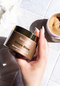 Choco Glow kroppshydrator i en cylindrisk brun behållare med ett gyllene lock. Texten inkluderar produktnamn och volym, placerad på en ljus yta.