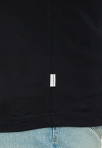 Schwarzes Baumwoll-T-Shirt mit einem seitlichen Etikett "ORIGINALS" und genähtem Saum, präsentiert über hellblauem Jeansstoff.