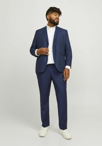 Costume bleu marine au design ajusté, composé d'une veste à simple boutonnage et de pantalon à devant plat, associé à une chemise blanche à boutons et des baskets blanches.