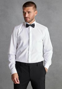 Cămașă albă cu cravată neagră, mâneci lungi, butoni negri și croi slim. Asortată cu pantaloni negri.