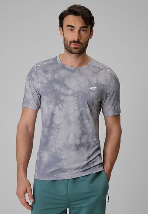 Mężczyzna o ciemnych włosach i brodzie, ubrany w szary krótkim rękawem koszulkę tie-dye i zielone spodnie, z jedną ręką w kieszeni na szarym tle.