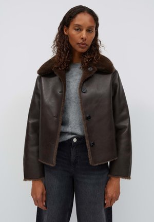 Woman wearing dark brown leather jacket with fur collar, gray sweater, and black jeans standing against a plain background.