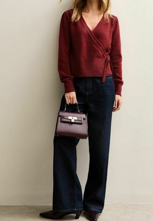 Kobieta ubrana w bordowy sweter kopertowy i ciemne jeansy z szerokimi nogawkami, trzymająca pasującą bordową torebkę i nosząca buty z noskiem w szpic.