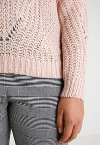 Pull en tricot rose clair avec un tissage lâche et un ourlet côtelé, associé à un pantalon à carreaux noir et blanc avec une coupe ajustée.