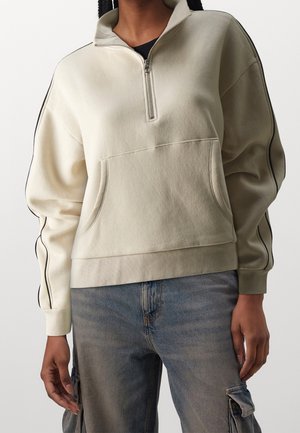 Persoon in een beige half-zip sweatshirt met zwarte streep op de mouwen en een voorzak, gecombineerd met vervaagde blauwe cargobroeken.