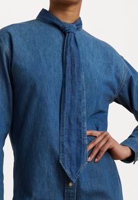 Camicia di denim con colletto annodato, caratterizzata da maniche lunghe e chiusura a bottoni. Il tessuto è di un blu medio con una texture liscia.