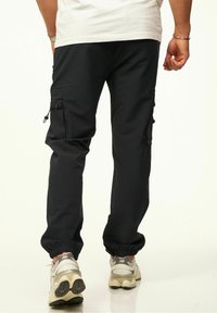 Schwarze Cargo-Hosen mit elastischen Bündchen, ausgestattet mit zwei Seitentaschen und einer glatten Textur. Kombiniert mit beigen Sneakers für den Kontrast.