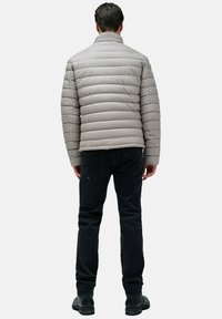 Chaqueta acolchada gris con patrón de acolchado horizontal, cuello alto y cierre de cremallera, combinada con jeans oscuros y zapatos negros.