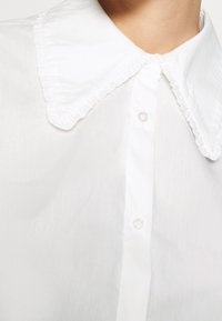 Chemise blanche à manches longues avec un grand col pointu orné de bords délicatement froncés, portée près du cou.