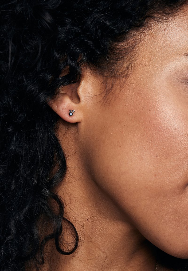Srebrne kolczyki stud z trzema małymi kamieniami szlachetnymi—dwoma okrągłymi niebieskimi i jednym przezroczystym—ustawione w kwiatowy wzór, noszone na lewym uchu.