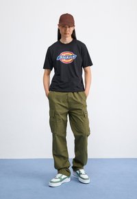 Svart grafisk T-skjorte med "Dickies"-logo, olivengrønne cargo-bukser og hvite sneakers med grønne detaljer. Brun caps fullfører antrekket.