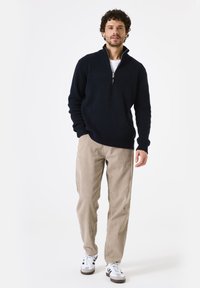 Marine blauwe half-ritskraag pullover in een gestructureerde brei, gecombineerd met licht beige corduroy broek. Model draagt witte sneakers met zwarte strepen.
