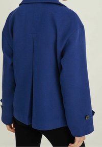 Blå ulljacka med ryggslits, sänkta axlar och knäppningar vid ärmarna. Den strukturerade designen ger en skräddarsydd look.