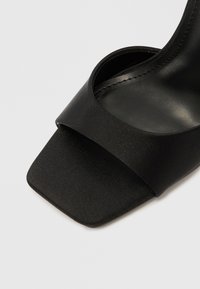 Μαύρο σατέν πέδιλο με τακούνι, με φαρδιά λωρίδα που διασχίζει τα δάχτυλα, τετράγωνο σχήμα στο μπροστινό μέρος και ομαλή, ματ υφή. Απλός σχεδιασμός συνολικά.