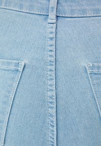 Tela de mezclilla azul claro con una costura visible en el centro. Presenta dos bolsillos traseros, cada uno con costuras blancas y esquinas angulares.