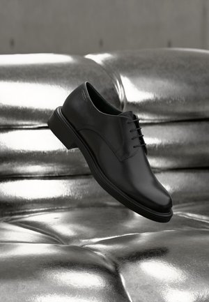 Crna kožna cipele s vezicama lebdi iznad sjajnog, srebrnog metalnog jastučića s gumbima na sofi.