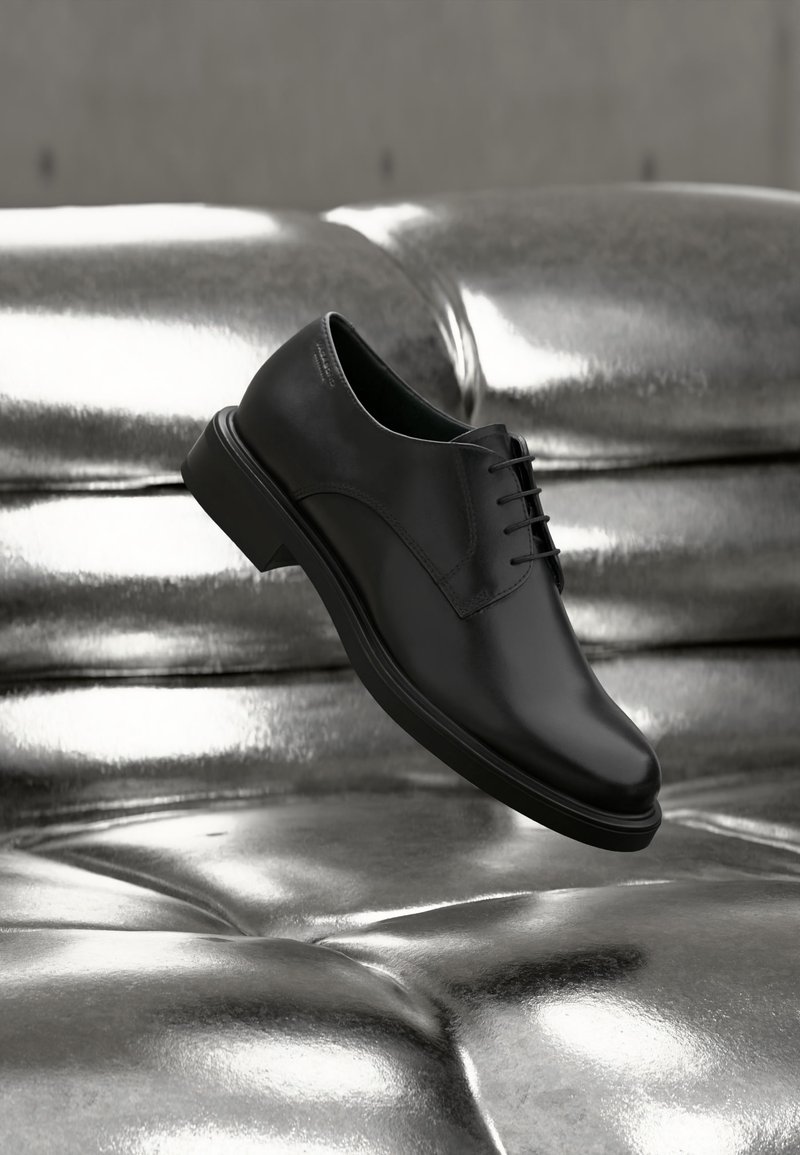 Μαύρο δερμάτινο δετό παπούτσι φορεσιάς που αιωρείται πάνω από ένα γυαλιστερό, ασημένιο μεταλλικό μαξιλάρι καναπέ με καπιτονέ.