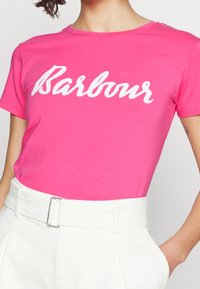 Růžové bavlněné tričko s bílým kurzívou napsaným logem "Barbour", kulatým výstřihem a krátkými rukávy. V kombinaci s bílými kalhotami s vysokým pasem.