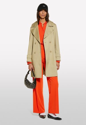 Beige, doppelt knöpfbarer Trenchcoat, orange Hemd und Hose, schwarze Mütze, schwarze Tasche mit Ösen, schwarze Ballerinas mit weißen Socken.