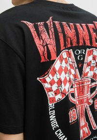 Sort t-shirt med korte ærmer, der har et sprækket rødt "VINNER"-grafik, ternede flag og et trofædesign. Teksten omfavner logoet.