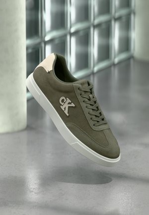 Olive green suede sneaker with white sole and CK logo, displayed against a gray floor and glass block wall background.