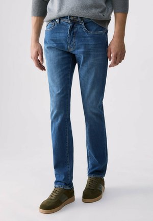 Homme portant un jean bleu, un pull gris et des baskets vertes, debout sur un fond clair, mains détendues le long du corps.