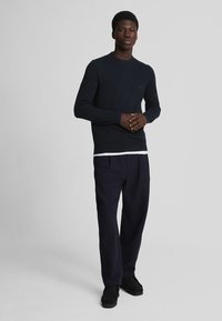 Suéter de punto azul marino con mangas largas, cuello redondo y un detalle sutil del logo. Combinado con pantalones oscuros de corte holgado y zapatos negros.