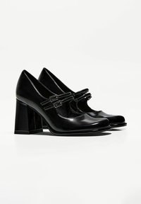 Black patent leather high-heeled shoes with a squared toe, large block heels, and two buckled straps across the front.