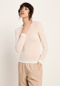 Frau mit kurzem, dunklem Haar, die ein enganliegendes, langärmliges, beige geripptes Oberteil und lockere, beige Cord-Hosen trägt, posiert mit einer Hand in der Tasche.