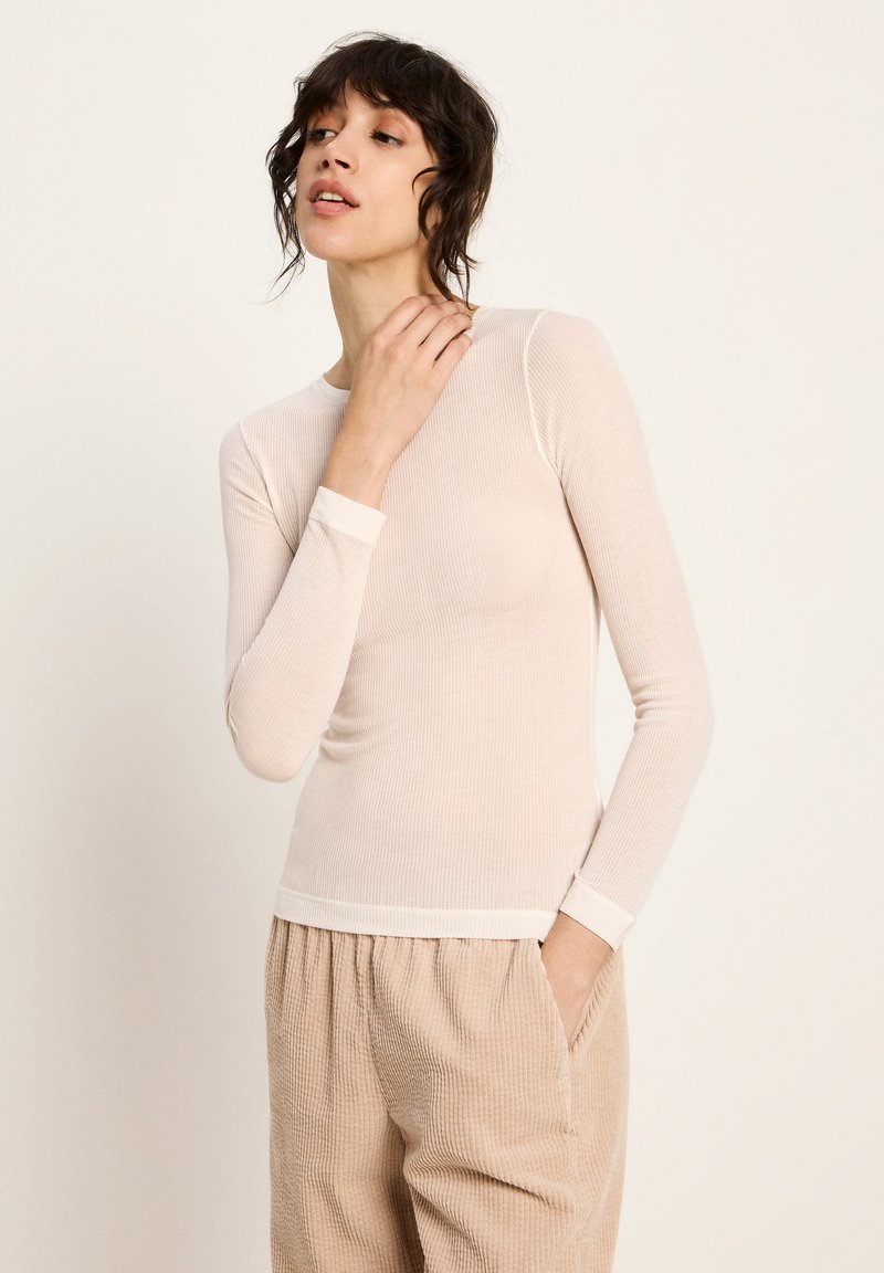 Frau mit kurzem, dunklem Haar, die ein enganliegendes, langärmliges, beige geripptes Oberteil und lockere, beige Cord-Hosen trägt, posiert mit einer Hand in der Tasche.