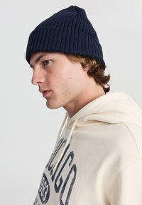 Berretto lavorato a maglia blu marina con texture a costine, cima appuntita, indossato con una felpa beige con una stampa grafica scura. Focus sulla testa e sulla parte superiore del corpo.