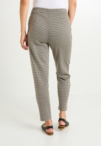 Femme portant un pantalon tapered checké beige et noir avec un haut blanc et des sandales plates noires, vue de dos sur fond blanc.