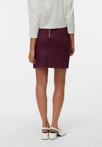Jupe mini bordeaux avec une texture lisse et un détail de fermeture éclair à l'arrière, associée à des chaussures à talons blanches et un haut léger ample.