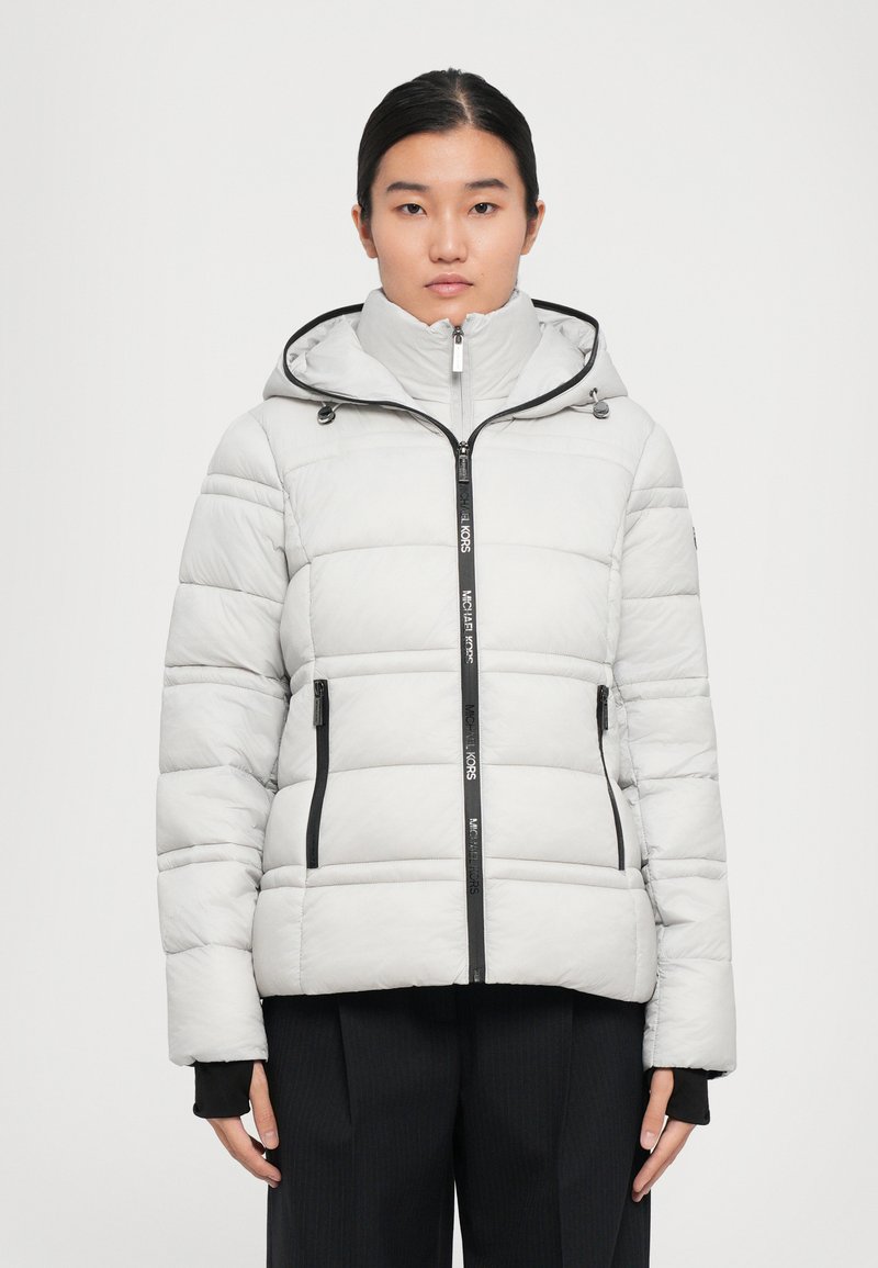 Femme portant une veste matelassée gris clair avec capuche, fermeture éclair sur le devant et poches zippées noires, associée à un pantalon noir.