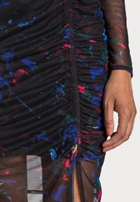 Zwarte jurk met kleurrijke spetters in blauw, rood en groen. Heeft ruches en een hoge split. Gemaakt van een doorschijnende, gestructureerde stof.