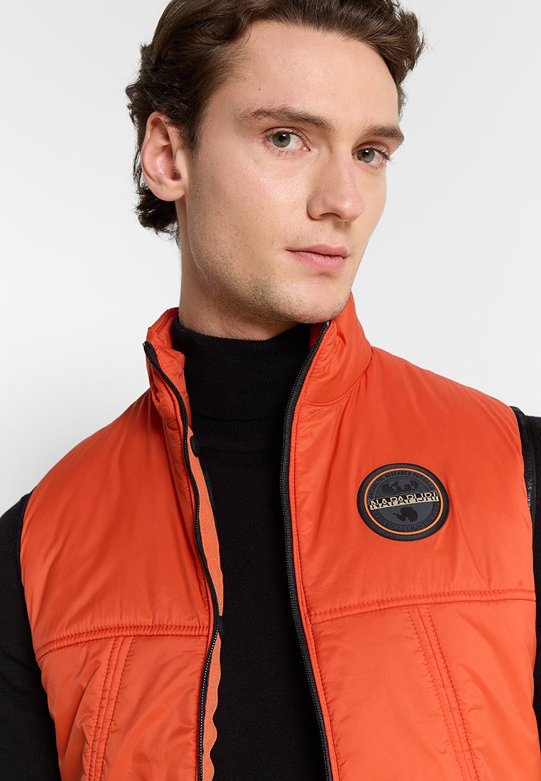 Lekka pomarańczowa kamizelka puchowa z czarnym zamkiem błyskawicznym i okrągłą naszywką z logo na piersi, z wysokim kołnierzem oraz teksturowanym materiałem.