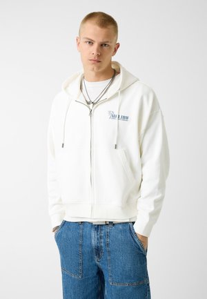 Giovane uomo con capelli corti che indossa una felpa con cappuccio bianca con cerniera e logo "Malibu" e jeans blu, in piedi con le mani in tasca su sfondo bianco.