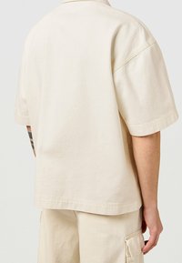 Camisa de manga corta de gran tamaño en algodón de color beige claro. Presenta un cuello clásico, corte holgado y mínimos detalles de costura.