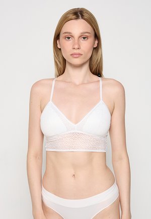 Młoda kobieta o prostych blond włosach, ubrana w biały koronkowy bralet i białą bezszwową bieliznę, stojąca na jednolitym jasnym tle.