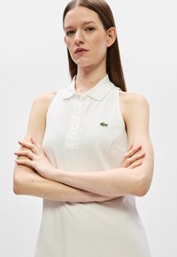 Jeune femme avec de longs cheveux bruns portant un polo blanc sans manche avec un petit logo de crocodile vert, bras croisés, fond neutre.