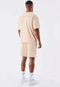 Completo di t-shirt e pantaloni corti in cotone color pesca. La t-shirt ha un logo stampato sul retro. Abbinato a calzini bianchi e sneakers.