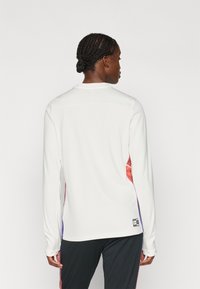 Dlouhý rukáv sportovní top v bílé látce s bočními panely v červené a fialové, s kulatým výstřihem a nášivkou loga na lemu.