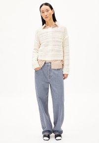 Cremfarbener, strukturierten Strickpullover über einem gestreiften beigen Hemd, kombiniert mit lockeren hellblauen Jeans und schwarzen Sandalen mit weißen Akzenten.