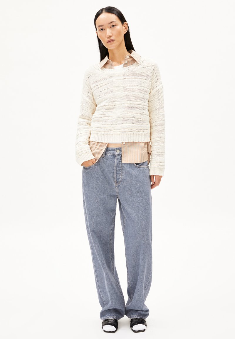 Cremfarbener, strukturierten Strickpullover über einem gestreiften beigen Hemd, kombiniert mit lockeren hellblauen Jeans und schwarzen Sandalen mit weißen Akzenten.